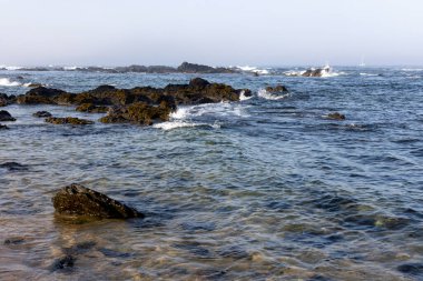 Görüntü, koyu renkli kayaların nispeten sakin, mavi-gri suya battığı bir sahneyi gösteriyor. Kayaların etrafında küçük dalgalar görülebilir. Gökyüzü bulutlu ve soluk gri-mavi. 