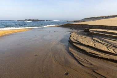 Görüntü, kıyı manzarasının geniş bir görüntüsünü gösteriyor. Ön planda koyu kahverengi, katmanlı kaya oluşumlarıyla görülebilir erozyon desenleri gösteren bir gelgit havuzu var..