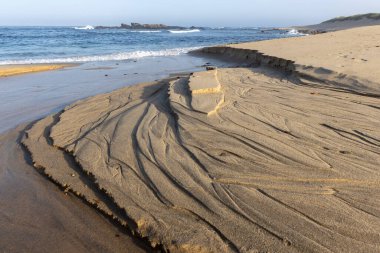 Kıyı kum oluşumunun yakın çekim görüntüsü koyu çizgilerle kaplı katmanlı bir dokuyu akıcı ve soyut bir desen oluşturur. Kum daha koyu, neredeyse siyah çizgilerle açık bej renkte görünüyor..