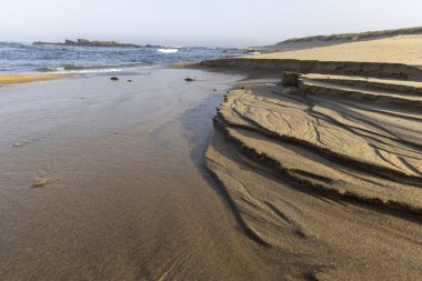 Görüntü, bir sahil kum oluşumunun yakın görüntüsünü sunuyor. Ön planda koyu kahverengi, katmanlı kaya yapısı görülebilir çizgiler ve kıvrımlarla eşsiz bir doku sergiliyor.