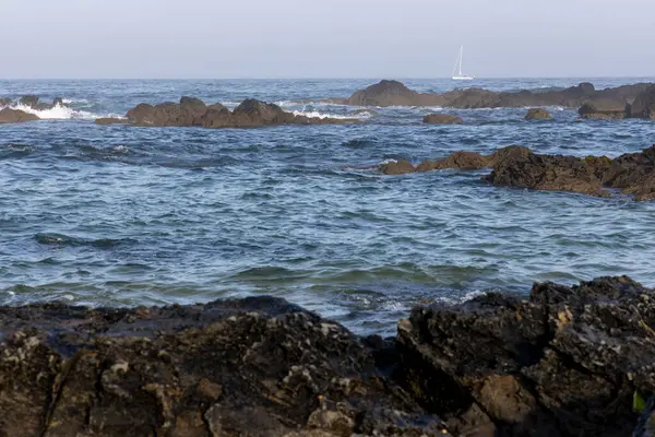 Görüntü, okyanus dalgalarıyla dolu kayalık bir kıyı şeridini betimliyor. Ön planda koyu renkli kayaların kısmen suya battığını gösteriyor. Orta yerde su yüzeyinde hareket eden küçük dalgalar var.
