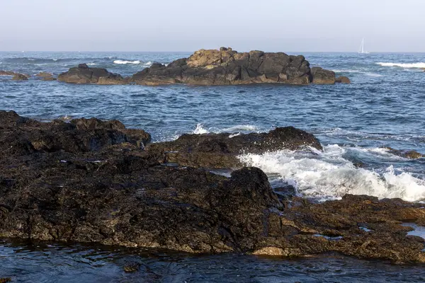 Görüntü, sudan çıkan sert kayaların olduğu bir kıyı sahnesini gösteriyor. Okyanus dalgaları görülebilir, kayalara çarptıkları yerde beyaz köpük görülebilir. Su koyu mavi..