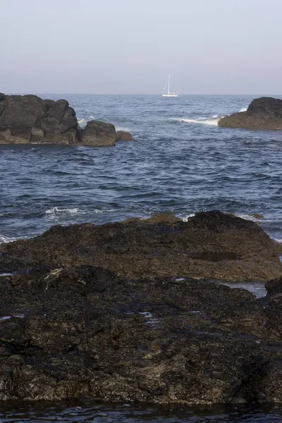 Görüntü, çalkantılı bir su kütlesinin kenarındaki karanlık ve pürüzlü kayaların yakın görüntüsünü gösteriyor. Su koyu mavi-gri renktedir, küçük ve orta büyüklükte dalgalar desenli bir yüzey oluşturur.. 