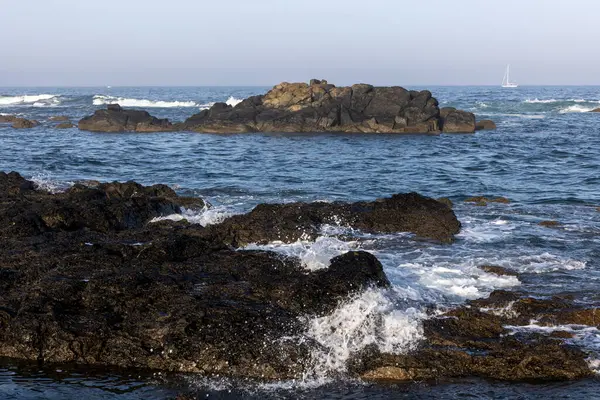 Görüntü, karanlık ve dalgalı kayaların hakim olduğu bir sahil sahnesini gösteriyor. Mavi su kütlesinden çıkan kayalar. Dalgalar kayalara çarpıp beyaz köpük oluşturuyor. Su biraz dalgalı görünüyor.. 