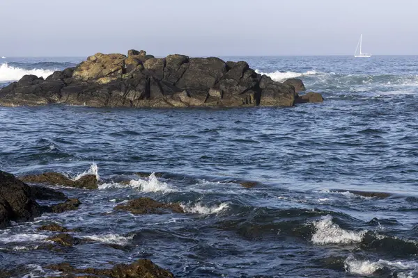 Görüntü, okyanustaki kayalıkların manzaraya yönelik bir fotoğrafını gösteriyor. Kayalar koyu renklidir, pürüzlü ve dokulu görünürler. Okyanus suyu görünür dalgaları olan koyu bir mavidir..