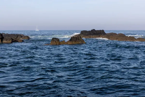 Görüntü, okyanus dalgalarıyla kayalık bir sahil şeridini gösteriyor. Su koyu mavi renktedir, dalgalarda beyaz noktalar görülebilir. Koyu renkli kayalar kısmen suya gömülüdür..