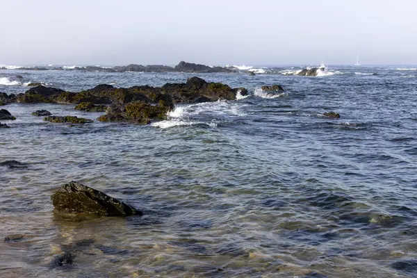 Görüntü, koyu renkli kayaların nispeten sakin, mavi-gri suya battığı bir sahneyi gösteriyor. Kayaların etrafında küçük dalgalar görülebilir. Gökyüzü bulutlu ve soluk gri-mavi. 