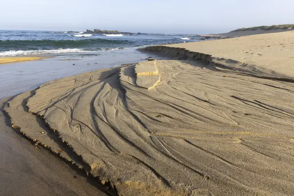 Karmaşık desenler ve dokular gösteren kıyı kum oluşumunun yakın görüntüsü. Kumun katmanlı ve çizgili yapısını vurgulayan açık bej ve kahverengi tonlara sahiptir..