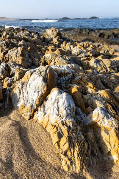 Sahilde kayaların yakın çekim görüntüsü. Kayalar esasen açık kahverengi ve bronzlaşmış, açık renkli çizgiler, neredeyse beyaz, renklendirilmiş. Doku pürüzlü ve düzensiz. Kayalar çeşitli şekiller gösterir.