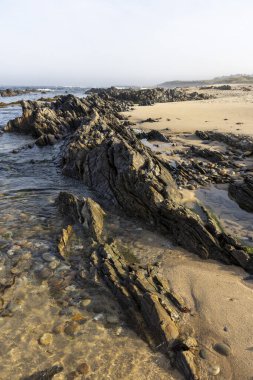 Yüksek açılı, koyu renkli bir kaya oluşumunun yakın çekim görüntüsü okyanustan kumlu bir sahile kadar uzanıyor. Kayalar pürüzlü, yıpranma ve aşınma belirtileri gösteriyor..  