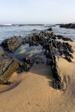 Yüksek açılı, dalgaların çekildiği kayalık bir kıyı şeridine yakın çekim. Görüntü koyu renkli, düzensiz şekilli kayaları gösteriyor. Bazıları ıslak, bazıları kuru. Açık renkli kum görünür..