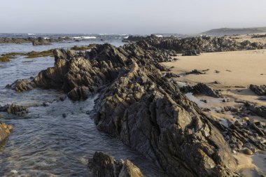 Görüntü, okyanustan kumlu bir sahile doğru uzanan belirgin koyu gri ve kahverengi bir kaya oluşumunun yer aldığı bir kıyı manzarasını gösteriyor. Kayalar şekil ve boyut olarak düzensiz..