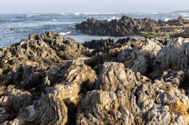 Görüntü kayalık bir sahil şeridini yakından gösteriyor. Kayalar ağırlıklı olarak koyu gri ve kahverengidir, değişik doku ve şekillere sahiptir. Bazı kayalarda yıpranma ve aşınma izleri var.. 