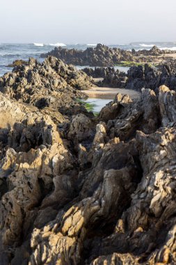 Yüksek açılı, kayalık bir kıyı şeridine yakın çekim. Görüntü koyu gri ve kahverengi kayalar arasında küçük kumlu bir sahile giden dar bir geçidi ve durgun bir su kütlesini gösteriyor.. 