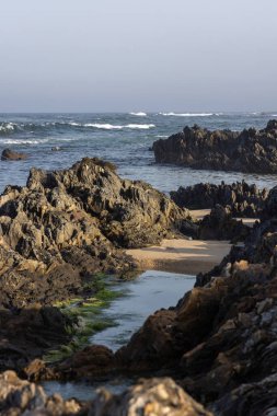Görüntü, bir sahil şeridindeki koyu, kaba desenli kayaların yakın görüntüsünü gösteriyor. Kayalar düzensiz şekillenmiş ve aşınma belirtileri gösteriyor. Okyanus dalgaları görünür..