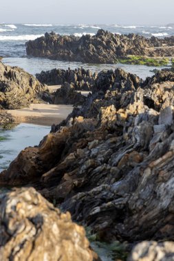 Görüntü kayalık bir sahil şeridini yakından gösteriyor. Kayalar kahverengi, gri ve bej tonlarıdır, kaba dokular, çeşitli şekiller ve boyutlar sergilerler.. 