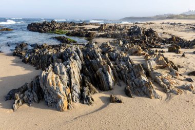 Görüntü bir plaj sahnesinin peyzaj odaklı bir fotoğrafını gösteriyor. Ön planda değişik boylarda ve şekillerde koyu gri ve siyah kayalar var..