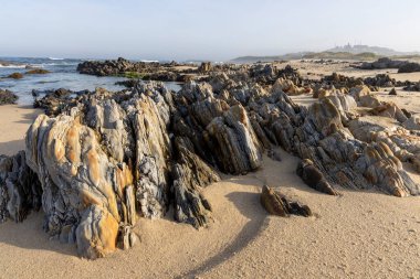 Görüntü, suyun kenarından uzanan sayısız katmanlı kaya oluşumlarıyla kumlu bir plajın panoramik bir manzara fotoğrafı. Kayalar ağırlıklı olarak gri ve kahverengi..