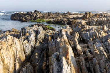 Görüntü, kıyı kayaları oluşumunun detaylı bir görüntüsünü gösteriyor. Kayalar ağırlıklı olarak gri ve bej renklidir, çeşitli doku ve şekiller sergilerler, bazıları katmanlı veya sütunlu görünür..  