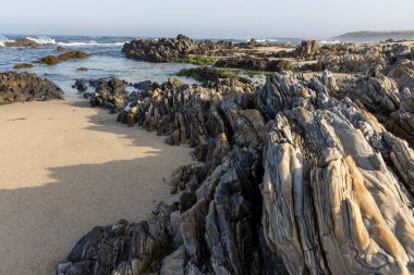 Görüntü dalgaların çekildiği kayalık bir kıyı şeridini gösteriyor. Ön planda koyu gri ve kahverengi katmanlı kayalar hakimdir, görülebilir çizgiler ve varyasyonlarla kaba bir doku sergilerler.