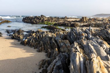Görüntü kayalık bir sahil şeridini gösteriyor. Değişen boyut ve şekillerden oluşan koyu renkli kayalar ön plana hükmedip okyanusa kadar uzanır..