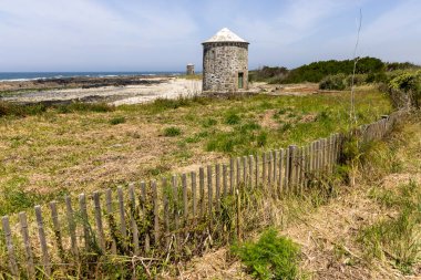 Görüntü, çimenli bir kum tepesinde yer alan eski, yıpranmış bir yel değirmeni kulesini tasvir ediyor. Basit bir ahşap çit ön planda çaprazlama ilerliyor.. 