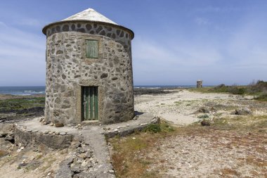 Görüntü, kıyı bölgesinde küçük yuvarlak bir taş kuleyi gösteriyor. Kule yaşlanmış ve yıpranmış görünüyor, konik bir çatısı var. Taş işçiliği sert ve düzensiz. Üzerinde küçük yeşil bir kapı görünür.