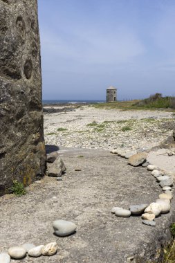 Görüntü, önplanda büyük, koyu renkli bir taş yapının yer aldığı ve manzarayı kısmen karartmış bir kıyı manzarasını gösteriyor. Orta zeminde bir çakıl taşı ve kumsal uzanır.