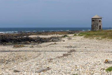Görüntü, ıssız bir kıyı manzarasının geniş bir görüntüsünü gösteriyor. Küçük, yıpranmış bir taş gözetleme kulesi kalıntısı, çakıllı bir plajın ortasında belirgin bir şekilde duruyor. Kumsal kayalık bir kıyı şeridine kadar uzanıyor.,