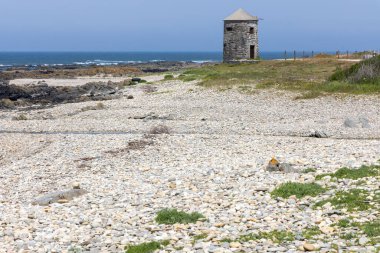 Görüntü, kıyı manzarasının geniş bir görüntüsünü gösteriyor. Ana odak noktası kayalık bir sahilde bulunan küçük, yıpranmış bir taş kule. Kule yaşlı ve muhtemelen terk edilmiş görünüyor. Kumsal sakin.