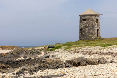 Görüntü kayalık bir sahil şeridinde yıpranmış bir taş kuleyi gösteriyor. Kule konik çatısı olan silindir şeklinde ve eski ve muhtemelen terk edilmiş görünüyor. Taşlar gri. .