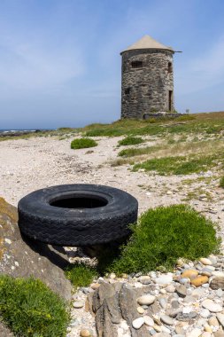 Görüntü, terk edilmiş, yıpranmış bir lastiğin çakıllı bir plajda yattığını gösteriyor. Lastik siyah ve yıpranmış görünüyor. Arka planda seyrek bitki örtüsü, düşük çalılıklar var.,