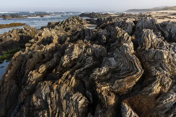 Yüksek açılı, kıyı kayaları oluşumuna yakın çekim. Görüntü, pürüzsüz ve pürüzsüz yüzeyler de dahil olmak üzere çeşitli dokulara sahip koyu kahverengi ve gri kayalardan oluşan karmaşık bir düzenleme gösteriyor.. 