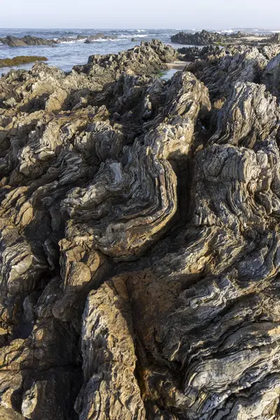 Karmaşık, girdap şeklinde dönen bir kaya yüzeyinin yakın plan fotoğrafı. Renkler ağırlıklı olarak koyu gri ve siyah, bej ve açık gri ipuçları var. Doku pürüzlü ve düzensiz..