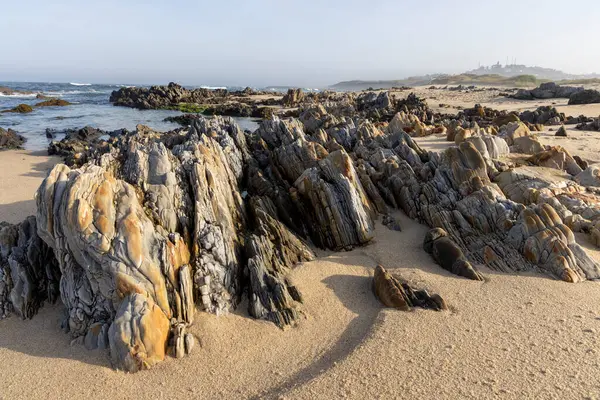Görüntü, suyun kenarından uzanan sayısız katmanlı kaya oluşumlarıyla kumlu bir plajın panoramik bir manzara fotoğrafı. Kayalar ağırlıklı olarak gri ve kahverengi..