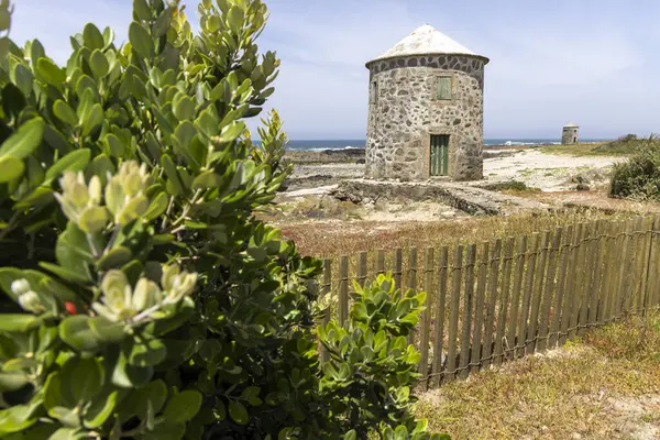 Görüntü, kıyı manzarasında bulunan yıpranmış bir yel değirmenini gösteriyor. Yel değirmeni konik bir kapakla silindirik..