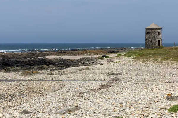 Görüntü, ıssız bir kıyı manzarasının geniş bir görüntüsünü gösteriyor. Küçük, yıpranmış bir taş gözetleme kulesi kalıntısı, çakıllı bir plajın ortasında belirgin bir şekilde duruyor. Kumsal kayalık bir kıyı şeridine kadar uzanıyor.,
