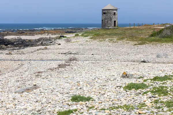 Görüntü, kıyı manzarasının geniş bir görüntüsünü gösteriyor. Ana odak noktası kayalık bir sahilde bulunan küçük, yıpranmış bir taş kule. Kule yaşlı ve muhtemelen terk edilmiş görünüyor. Kumsal sakin.