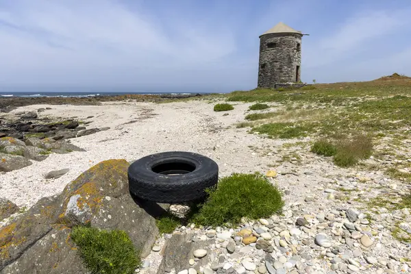Görüntü, arka planda terk edilmiş bir taş kule içeren kıyı manzarasını gösteriyor. Ön planda, ıskartaya çıkmış siyah bir lastik kayaların ve seyrek bitkilerin arasında kumlu zeminde duruyor.. 