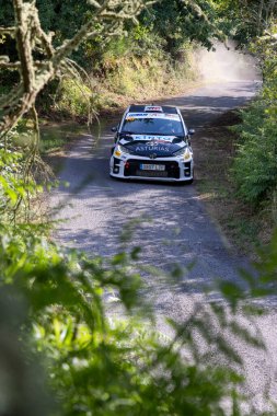Ourense, İspanya, 13 Haziran 2025: Görüntü, yemyeşil yeşilliklerin ortasında dar, dolambaçlı bir asfalt yolda ilerleyen beyaz rali aracının yakın görüntüsünü gösteriyor.