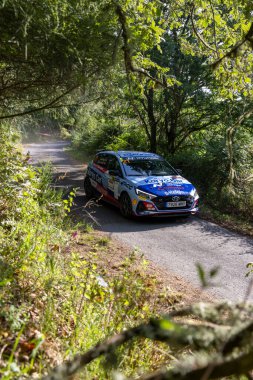 Görüntü, yemyeşil bir ormanda dar ve dolambaçlı bir yolda ilerleyen mavi ve beyaz bir rali arabasını gösteriyor..
