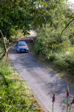 Ourense, İspanya, 13 Haziran 2025: yemyeşil bitki örtüsünü kesen dar, dolambaçlı bir yolda mavi bir rali aracının uzun açılı görüntüsü..