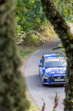 Ourense, İspanya, 13 Haziran 2025: Görüntü, ağaç dalları ve yapraklarından oluşan doğal bir çerçeveden alçak açıyla çekilen mavi bir rali arabasını gösteriyor..