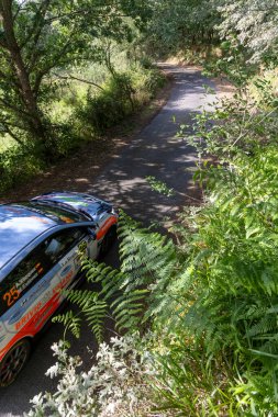 Ourense, İspanya, 13 Haziran 2025: Dar, dolambaçlı bir yolda, yemyeşil yeşilliklerle kısmen gizlenmiş gümüş bir rali aracının yüksek açılı yakın çekimi.