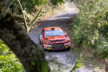 Ourense, İspanya, 13 Haziran 2025: Dar ve asfaltsız bir dağ yolunda keskin bir dönüş yapan turuncu renkli bir rali aracının yüksek açılı, hafif hafif anahtarlı görüntüsü.