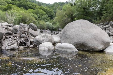 Görüntü, öncelikli olarak çeşitli büyüklükteki kaya ve kayalardan oluşan bir nehir yatağının yüksek açılı orta açılı görüntüsünü sunuyor. En büyük kaya gövdesi çerçevenin sağ merkezini kaplar ve pürüzsüz bir şekilde...,