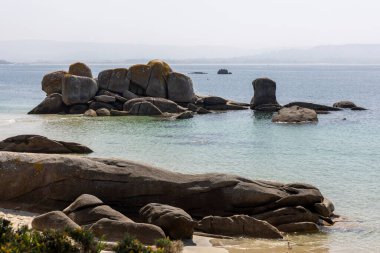 Arka planda büyük bir su kütlesi olan kayalık bir kıyı şeridi. Kıyı boyunca dağılan kayalar engebeli ve doğal bir manzara yaratıyor. Su sakin görünüyor..