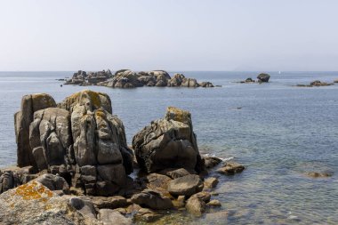 Arka planda büyük bir su kütlesi olan kayalık bir kıyı şeridi. Kıyı boyunca dağılan kayalar engebeli ve doğal bir manzara yaratıyor. Su sakin görünüyor..