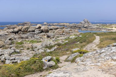 Suya giden yolu olan kayalık bir sahil. Arka planda bir yelkenli var. Su sakin..