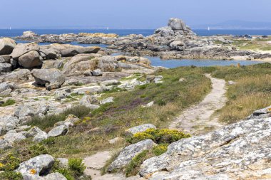 Suya giden yolu olan kayalık bir sahil. Arka planda bir yelkenli var. Su sakin..